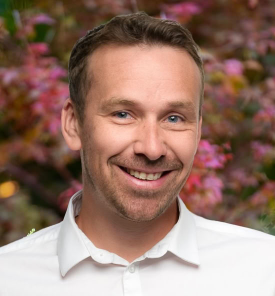 Quentin Hamet souriant portant une chemise blanche, avec un fond flou de feuillage coloré.