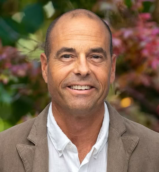 Alain Richner souriant portant une veste marron et une chemise blanche, avec un fond flou de feuillage coloré.