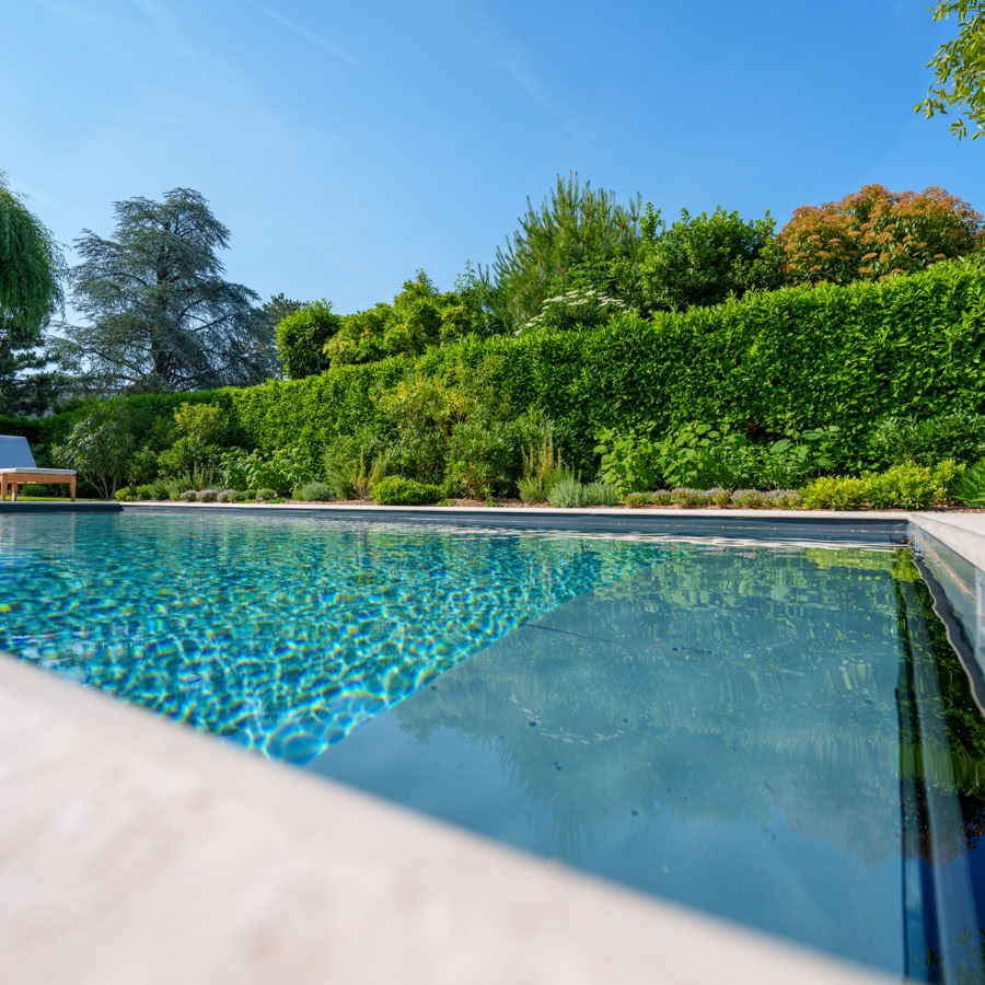 Piscine extérieure entourée de verdure avec des chaises longues blanches à gauche, ciel bleu en arrière-plan.