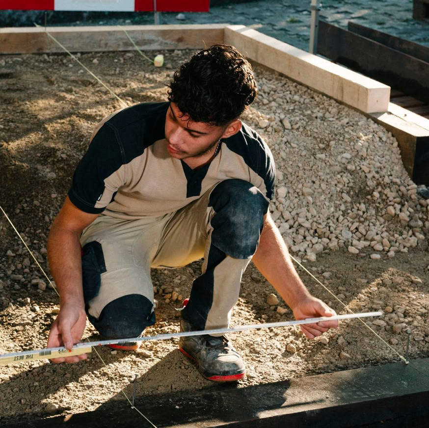 Apprenti Pottu & Seitz accroupi mesure une surface de terre avec un outil de nivellement.