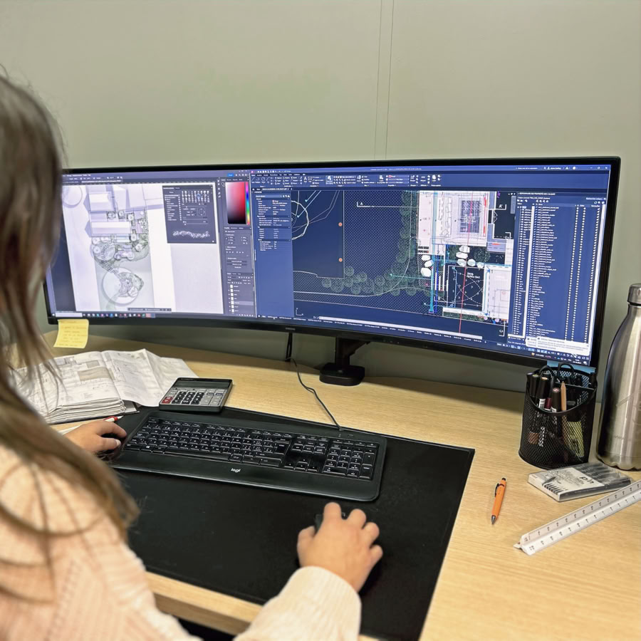 Architecte paysagiste qui travaille sur un ordinateur avec un écran large affichant des plans techniques. Un clavier et une souris sont sur le bureau, ainsi qu'un carnet, un téléphone, une règle et une bouteille.