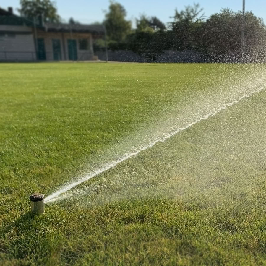 Spécialiste arrosage et robot. Arroseur automatique arrosant une pelouse verte avec des arbres et un bâtiment en arrière-plan.
