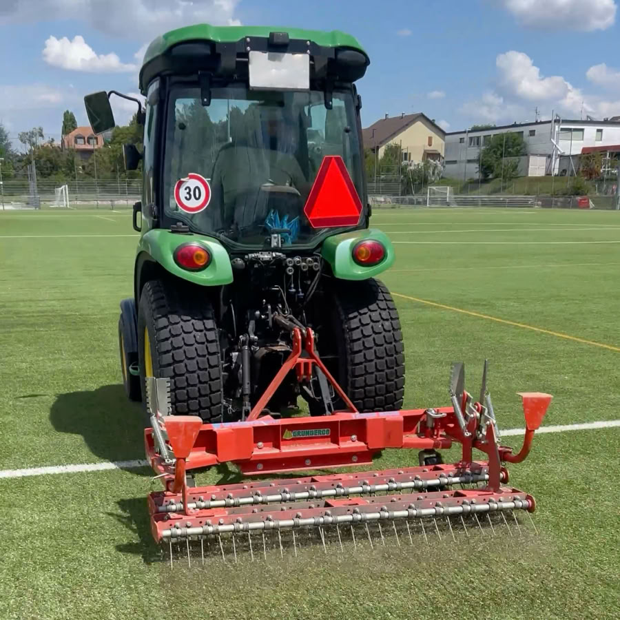 Tracteur vert avec un équipement rouge à l'arrière sur un terrain de sport en herbe synthétique, avec des bâtiments en arrière-plan.