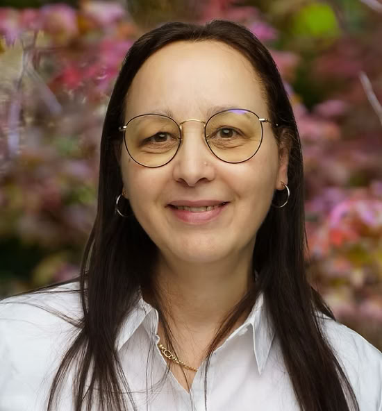 Fatiha Baaziz souriant portant une chemise blanche, avec un fond flou de feuillage coloré.