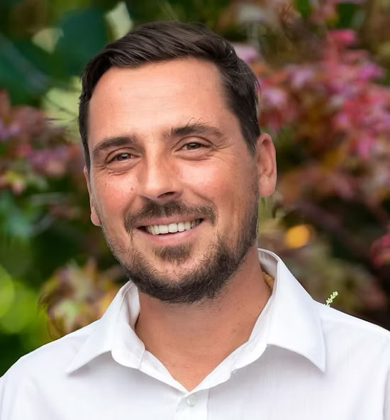 Jérémy Layat souriant portant une chemise blanche, avec un fond flou de feuillage coloré.