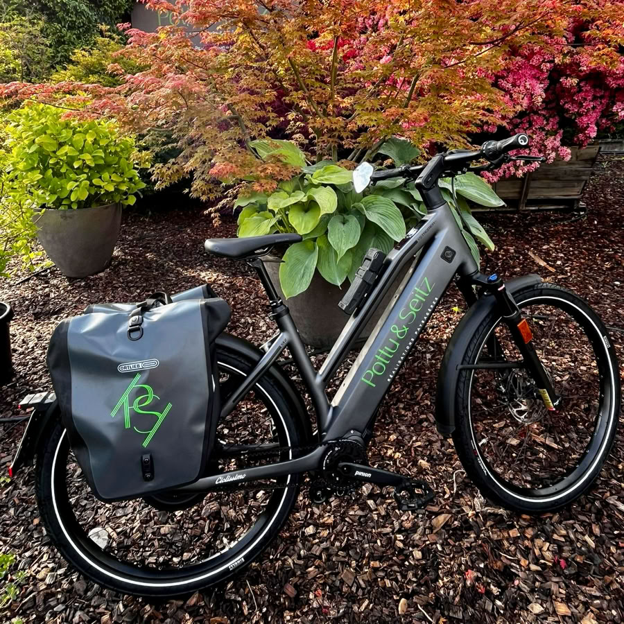 Vélo électrique Pottu & Seitz avec sacoche, posé sur un sol de gravier, entouré de plantes et d'arbustes aux feuilles colorées.