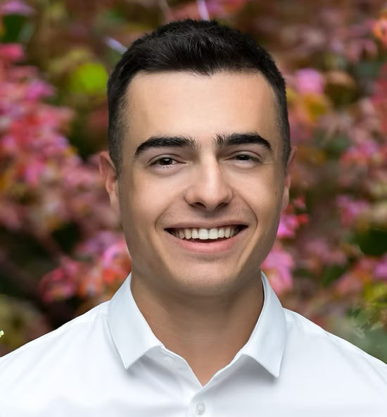 Romain Richner souriant portant une chemise blanche, avec un fond flou de feuillage coloré.