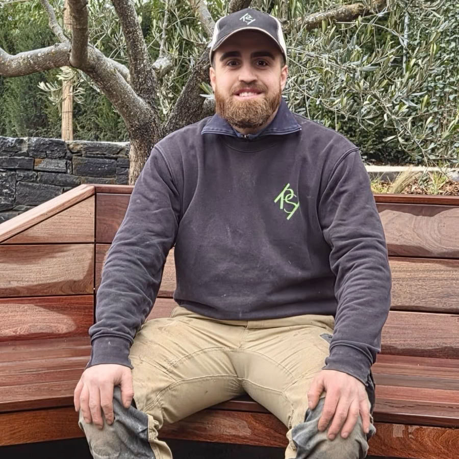 Daniel, paysagiste chez Pottu & Seitz, assis sur un banc en bois, portant une casquette, un pull foncé et un pantalon beige, avec des arbres en arrière-plan.