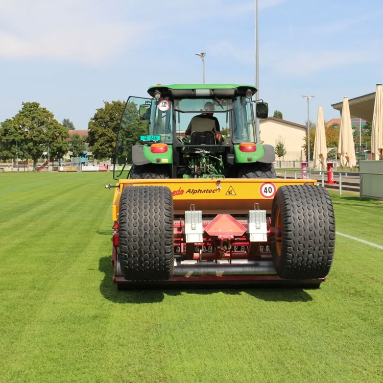 Tracteur vert sur un terrain de sport en herbe, avec un bâtiment à droite et des arbres en arrière-plan.