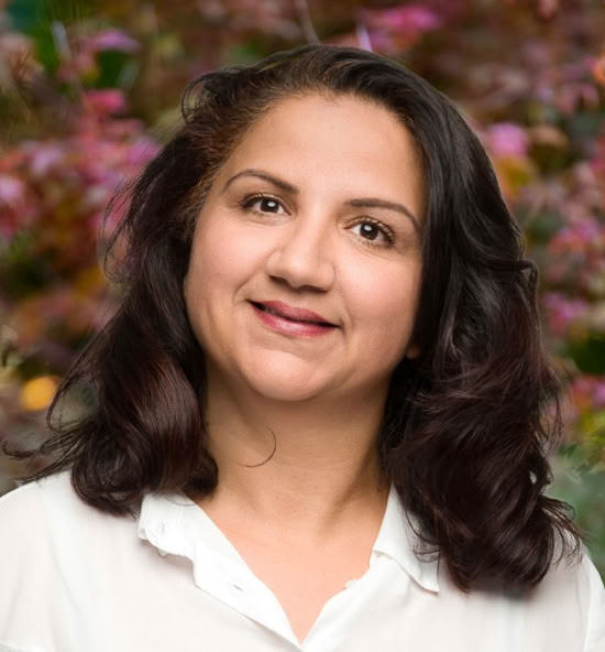 Radhia Anquetil souriant portant une chemise blanche, avec un fond flou de feuillage coloré.