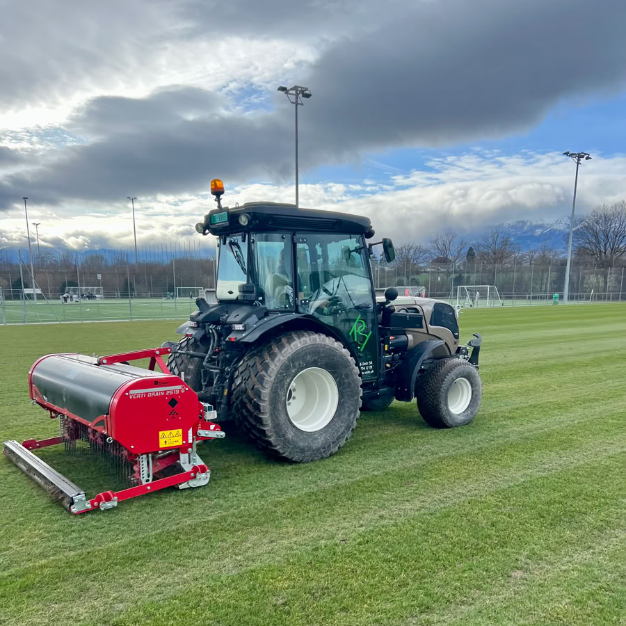 Réalisations sportives genève et vaud. Tracteur Pottu & Seitz avec un vertidrain sur un terrain de sport en herbe, sous un ciel nuageux.