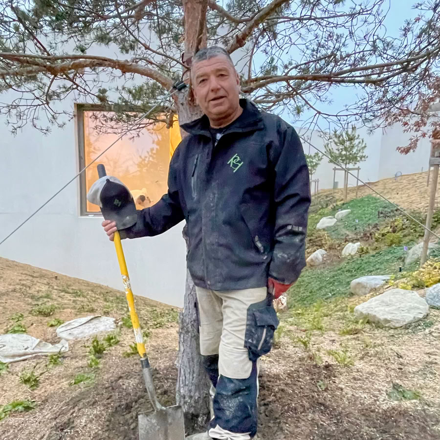 Thierry chef d'équipe chez Pottu & Seitz debout tenant une pelle, vêtu d'une veste noire et de bottes blanches, se trouve devant un arbre avec un panneau en arrière-plan.