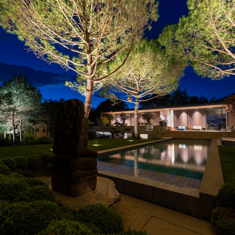 Jardin éclairé la nuit avec des arbres, une piscine rectangulaire et une maison moderne en arrière-plan.