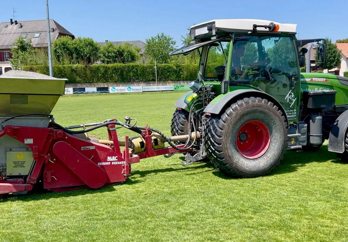 Tracteur vert avec une remorque rouge sur un terrain de sport en herbe, maisons et arbres en arrière-plan.