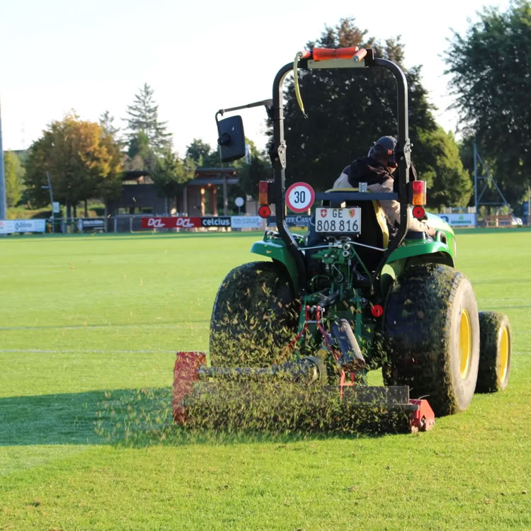 Tracteur vert tondant une pelouse sur un terrain de sport, avec des arbres en arrière-plan.