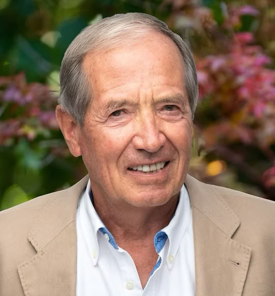 Jean-Jacques Seitz souriant portant une veste marron et une chemise blanche, avec un fond flou de feuillage coloré.