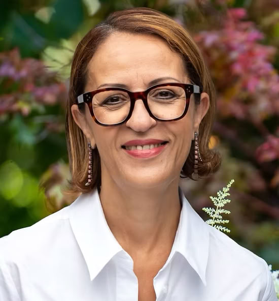 Touta Hadjadj souriant portant une chemise blanche, avec un fond flou de feuillage coloré.